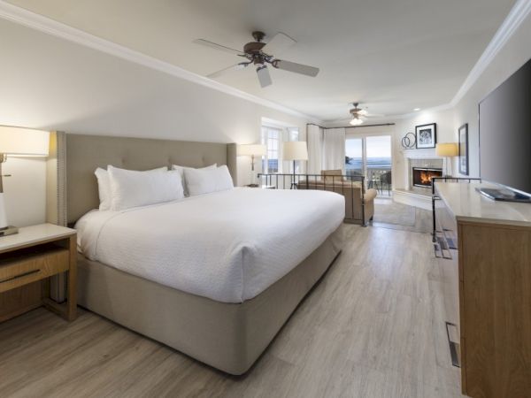 A spacious hotel room with a large king bed, bedside tables, seating area, desk, and a balcony view, all in light, neutral tones.