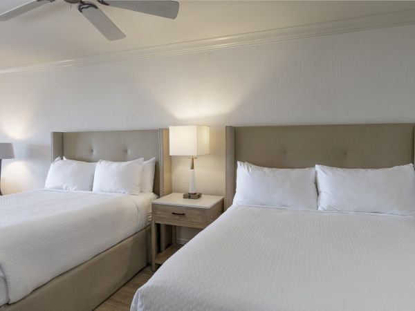 Two neatly made hotel rooms with two queen beds, matching nightstands, and lamps, under a ceiling fan.