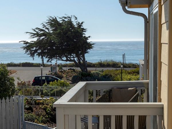 A balcony view overlooking a fence, trees, and the ocean beyond, with a clear sky and a side of a building visible.