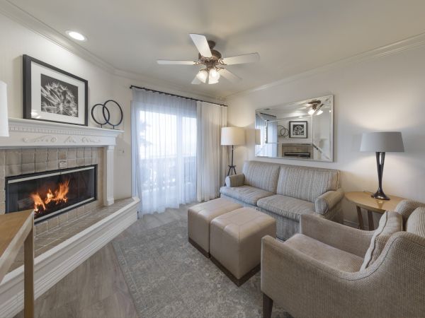 A cozy living room with a lit fireplace, beige recliners, a sofa, ottomans, lamps, and a large mirror, creating a warm, inviting space.