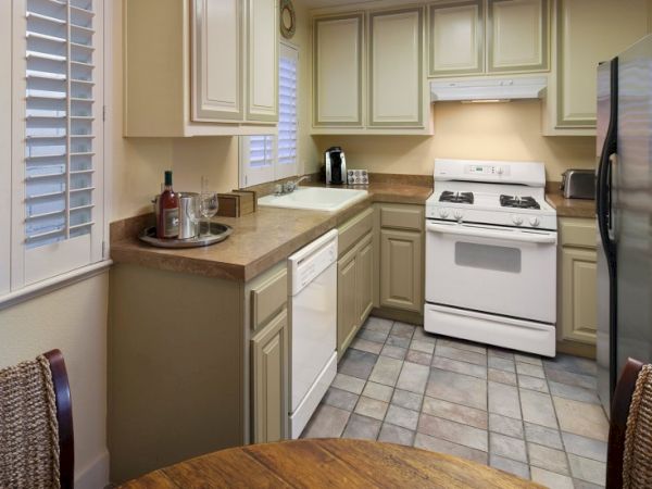 A compact L-shaped kitchen with beige cabinets, a white stove, stainless fridge, small counter space, a round table at the foreground, and a window with blinds.