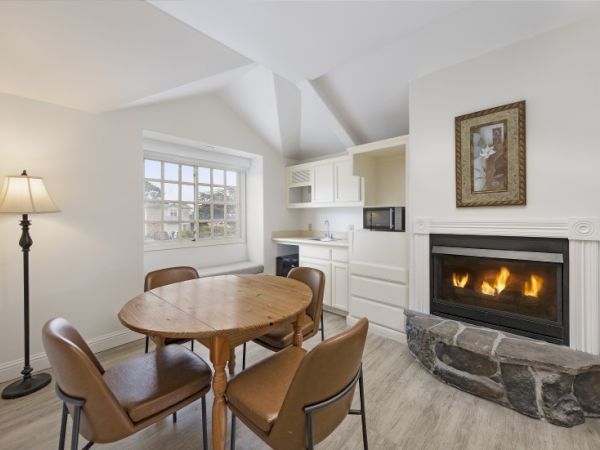 Cozy kitchenette with a round wooden dining table and four brown chairs, white cabinetry, a built-in fireplace with stone mantle, and a bright window letting in light.