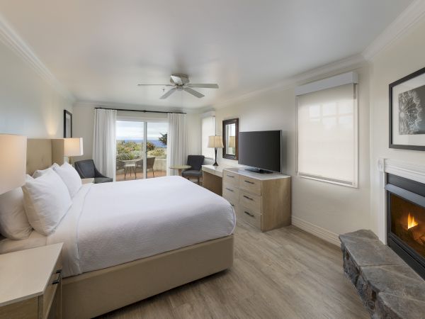 A bright hotel room with a king bed, nightstands, desk, dresser, flat-screen TV, fireplace, and a balcony view beyond sheer curtains.