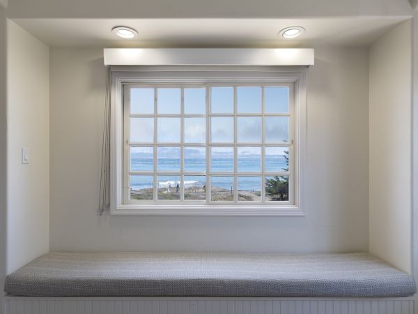 A cozy window nook with a large grid window, sea view outside, soft gray bench cushion, and recessed lighting above.