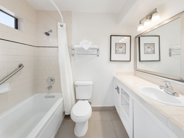 A clean, white bathroom with a bathtub/shower, a toilet, a long vanity with double sinks, a large mirror, and framed art above the vanity.