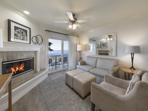 Cozy living room with a gas fireplace, beige sofa and chairs, ottomans, lamps, a mirrored wall, and a balcony view of the water outside.