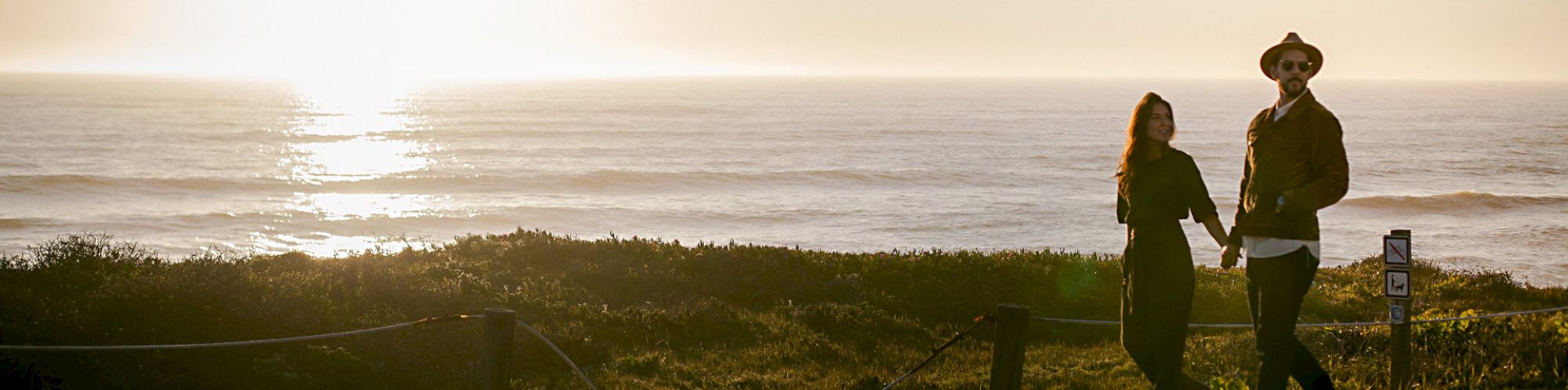 A couple walks hand in hand along a coastal path at sunset, the ocean shimmering ahead and warm light bathing the scene.