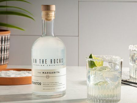A bottle labeled &ldquo;On the Rocks&rdquo; sits on a marble counter with two short glass drinks over ice, lime wedges, and a bowl of green apples in the background.