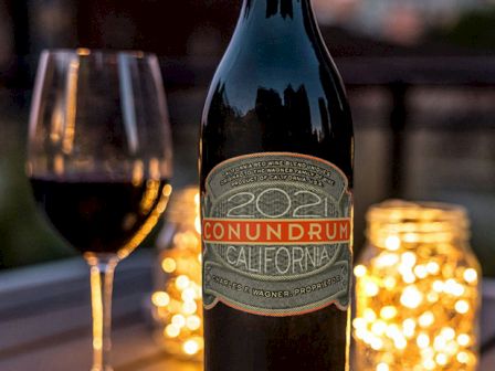 A bottle of wine with a glass, lit candles, and a warm outdoor dinner setup on a wooden table. The label reads "Conundrum California" with a glass of red wine beside it.