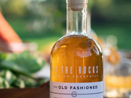 A stylish bottle of The Rocks Old Fashioned cocktail, set on a sunny outdoor table with greens and glasses in the background.