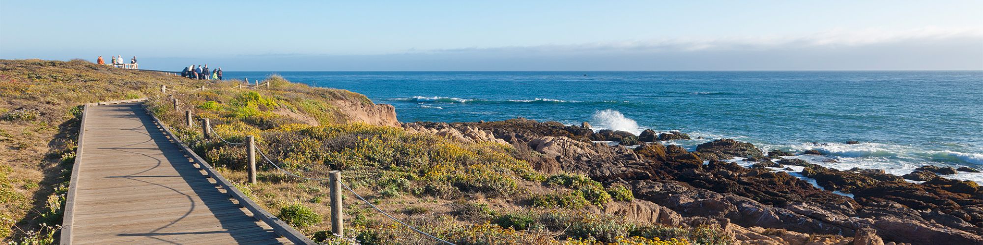 People gather near a coastal path that runs along rocky shorelines; waves crash softly as the ocean stretches to the horizon.