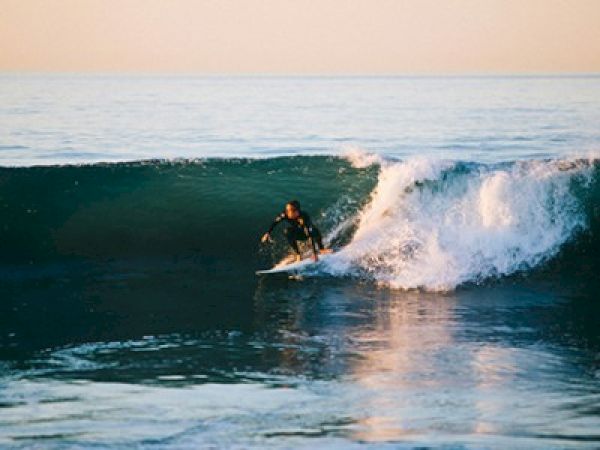 A surfer balances on a board, riding a small wave with a dark wetsuit as the sunlit sea glistens in the background.