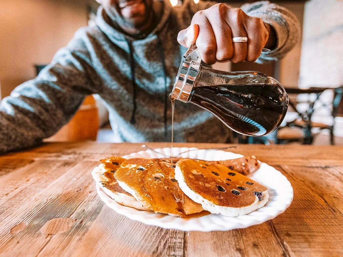 Person drizzles syrup over pancakes on a wooden table, pouring from a small pitcher; cozy, casual breakfast setup.