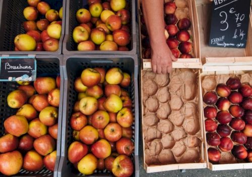 A market stall with crates of apples and pears on the left, and a person handling a box of small apples on the right, with price boards.
