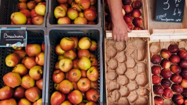 A market stall with crates of apples and pears on the left, and a person handling a box of small apples on the right, with price boards.