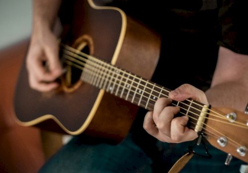 A person playing an acoustic guitar, close-up on fingers fretting the neck and strumming the strings on a dark guitar.