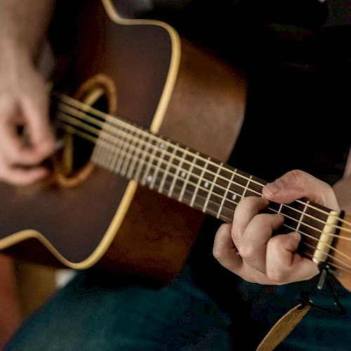 A person playing an acoustic guitar, close-up on fingers fretting the neck and strumming the strings on a dark guitar.