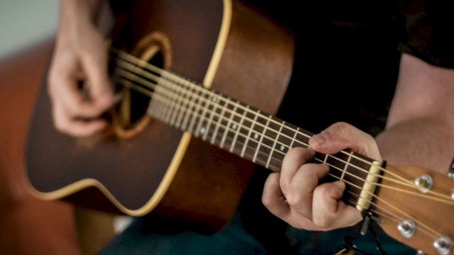 A person playing an acoustic guitar, close-up on fingers fretting the neck and strumming the strings on a dark guitar.