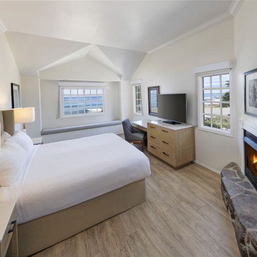 A bright hotel room with a king bed, nightstands, a desk, a dresser, a wall-mounted TV, and a fireplace beside a large windowed nook.