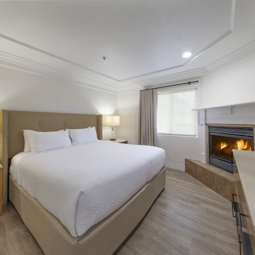 A modern hotel room with a king bed, bedside lamps, a desk and chair, a fireplace, and a TV, all in neutral tones, cozy and bright.