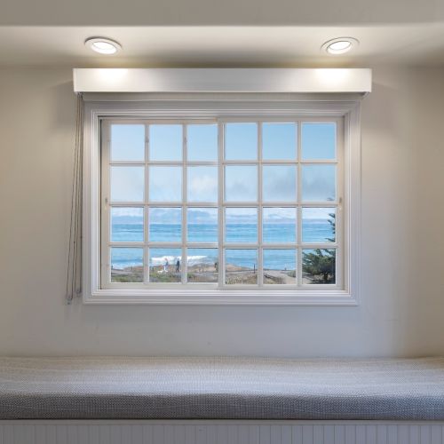 A cozy window seat nook with a large grid window overlooking the ocean, recessed lighting, and a cushioned bench beneath it.