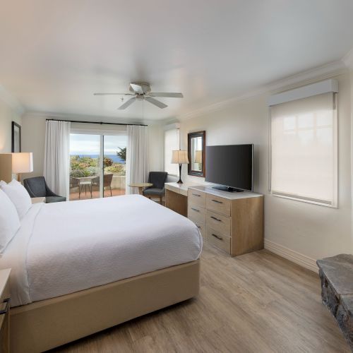 A bright hotel room with a king bed, nightstands, a dresser with a TV, a fireplace, and a sliding door to a balcony overlooking a landscape.