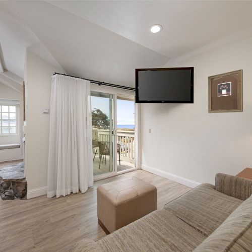 A bright living area with a beige sofa facing a wall-mounted TV, a small ottoman, a sliding door to a balcony, and a dining nook in the background, under soft recessed lighting.