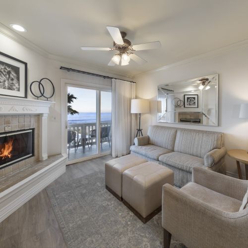 Cozy living room with a lit fireplace, light-gray furniture, a large mirror, sliding door to a balcony, and ocean view in the distance, bright and inviting.