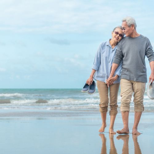 A senior couple walks barefoot along the beach, holding hands, smiling, and carrying their shoes as waves wash over the shore.