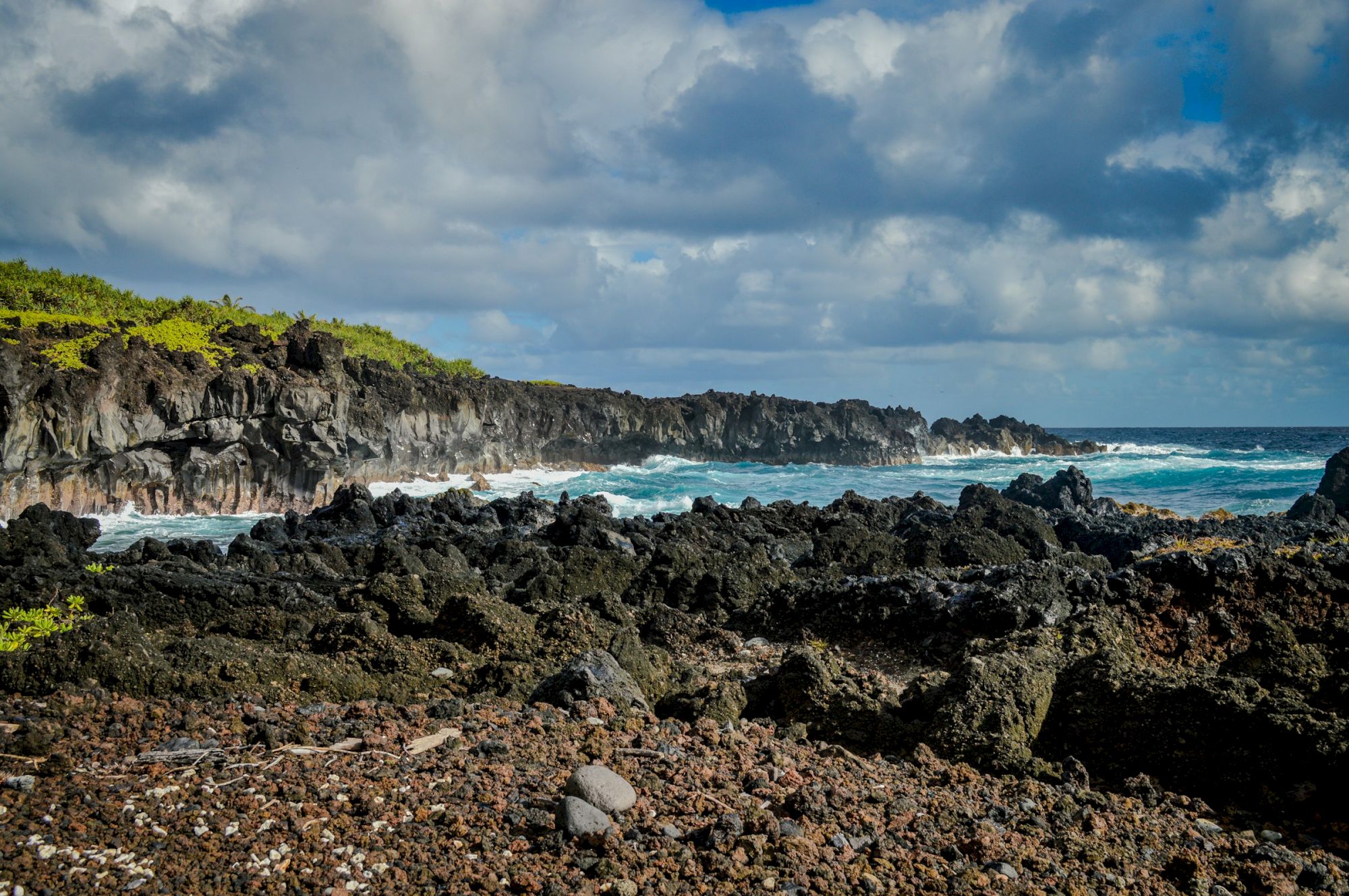 A rocky coastal scene with dark volcanic rocks, a pebbly shore, green shrub-covered cliffs, and blue ocean waves under a partly cloudy sky.